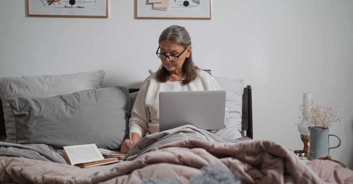 A woman with grey hair and glasses works on a laptop in bed, writing notes, with an open book beside her.
