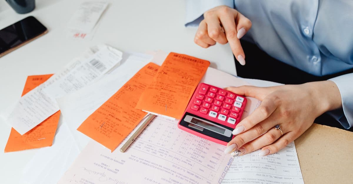 A woman checking a pile of orange and white receipts against a printed ledger, one hand on a pink calculator.