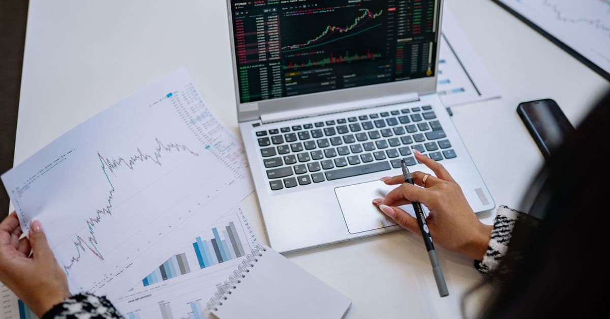 A woman at a white desk holding printed line and bar charts beside a laptop showing market charts, with a notebook and a pen.