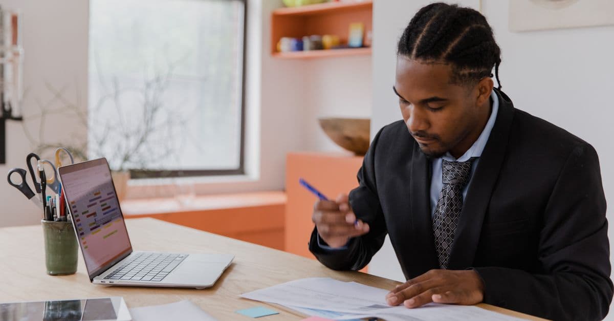 A man in a suit reads printed financial statements at a desk, pen in hand, beside a laptop showing a colourful spreadsheet.